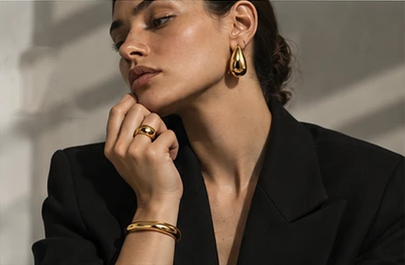 Mulher de blazer preto com brinco em gota, anel e pulseira dourados